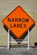 A bright orange warning sign with bold black text reading 'NARROW LANES' is mounted on a white frame structure against a beige brick wall.