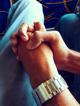 Two hands are clasped together in a gesture of unity or partnership. One person wears a silver metal watch on their wrist. The background includes the seat of a vehicle with a blue fabric and yellow patterns.
