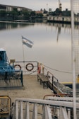A docked boat with an Argentine flag waving on the stern. The boat has lifebuoys and benches on its deck. In the background, there's a calm body of water reflecting the muted sky and waterfront buildings. Trees and an industrial structure with a crane are visible along the shore.