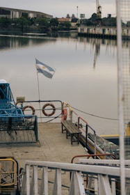 A docked boat with an Argentine flag waving on the stern. The boat has lifebuoys and benches on its deck. In the background, there's a calm body of water reflecting the muted sky and waterfront buildings. Trees and an industrial structure with a crane are visible along the shore.