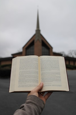 An open book is being held up outdoors with a church in the background. The church features a prominent steeple and a triangular roof. The sky is overcast, lending a gray hue to the scene. The person holding the book is wearing a brown sweater.