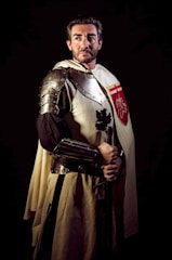 A person dressed as a medieval knight, wearing metal armor and a cloak with a heraldic symbol. The setting is dramatic with dark lighting, highlighting the armor and the individual's serious expression.