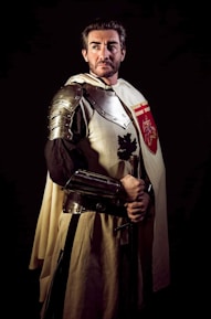 A person dressed as a medieval knight, wearing metal armor and a cloak with a heraldic symbol. The setting is dramatic with dark lighting, highlighting the armor and the individual's serious expression.