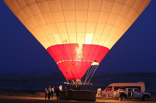 A large hot air balloon illuminated from within against a dark blue twilight sky. Several people are gathered around the basket preparing for a flight, and a vehicle is parked nearby, open and seemingly part of the launch setup.