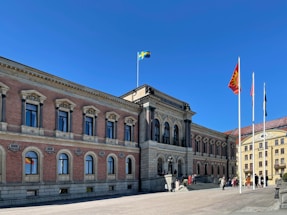 A large, classical-style building with arched windows and ornate architectural details stands under a clear blue sky. Several flags, including a Swedish flag, are hoisted on flagpoles in front of the building. People are gathered near the entrance, suggesting an event or gathering.