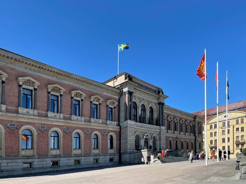 A large, classical-style building with arched windows and ornate architectural details stands under a clear blue sky. Several flags, including a Swedish flag, are hoisted on flagpoles in front of the building. People are gathered near the entrance, suggesting an event or gathering.