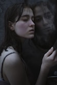 A person with closed eyes rests their head against a reflective surface, possibly a mirror, creating a soft, intimate moment. The lighting is dim, accentuating the wet hair and subtle textures of the skin.