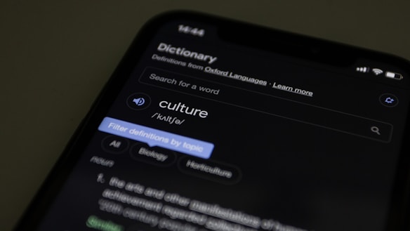 A smartphone displaying an online dictionary entry for the word 'culture'. The screen shows pronunciation, category tags such as Biology and Horticulture, and a part of the definition. The background is dark, focusing attention on the illuminated phone screen.