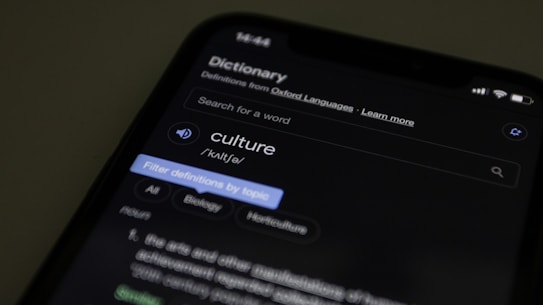 A smartphone displaying an online dictionary entry for the word 'culture'. The screen shows pronunciation, category tags such as Biology and Horticulture, and a part of the definition. The background is dark, focusing attention on the illuminated phone screen.
