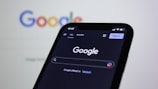 A smartphone displaying the Google search homepage in dark mode, with the Google logo visible on a blurred computer screen in the background.