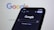 A smartphone displaying the Google search homepage in dark mode, with the Google logo visible on a blurred computer screen in the background.