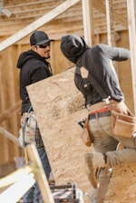 Two construction workers wearing hoodies and tool belts are actively engaged in work at a construction site. One worker is holding a large plywood board, while the other appears to be securing or inspecting structural framing. The background shows unfinished wooden frames, indicating ongoing construction.