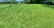 A lush, well-maintained green lawn stretches out with a wooden fence along one side. Trees and bushes border the grass, and the sky is visible in the background. A towel hangs on the fence, adding a hint of human presence.