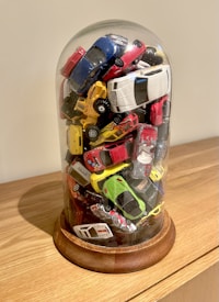 A collection of small toy cars stacked haphazardly inside a clear glass dome with a wooden base, placed on a light wooden surface. The toy cars are of various colors including red, blue, white, yellow, and green.