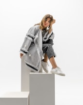 A woman is sitting on a set of minimalist white blocks, dressed in a stylish gray fringed coat and dark pants. She has a relaxed pose with her head resting on one hand and is wearing white sneakers. The background is a clean white, enhancing the modern and chic aesthetic of the image.