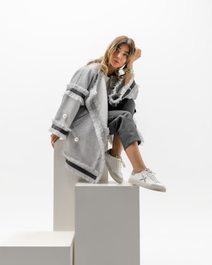 A woman is sitting on a set of minimalist white blocks, dressed in a stylish gray fringed coat and dark pants. She has a relaxed pose with her head resting on one hand and is wearing white sneakers. The background is a clean white, enhancing the modern and chic aesthetic of the image.