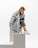 A woman is sitting on a set of minimalist white blocks, dressed in a stylish gray fringed coat and dark pants. She has a relaxed pose with her head resting on one hand and is wearing white sneakers. The background is a clean white, enhancing the modern and chic aesthetic of the image.