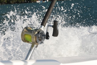 A close-up of a fishing reel mounted on a fishing rod, with a vibrant green line. The background features splashing water, suggesting movement and action, possibly from a boat cutting through the waves.