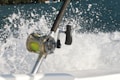 A close-up of a fishing reel mounted on a fishing rod, with a vibrant green line. The background features splashing water, suggesting movement and action, possibly from a boat cutting through the waves.