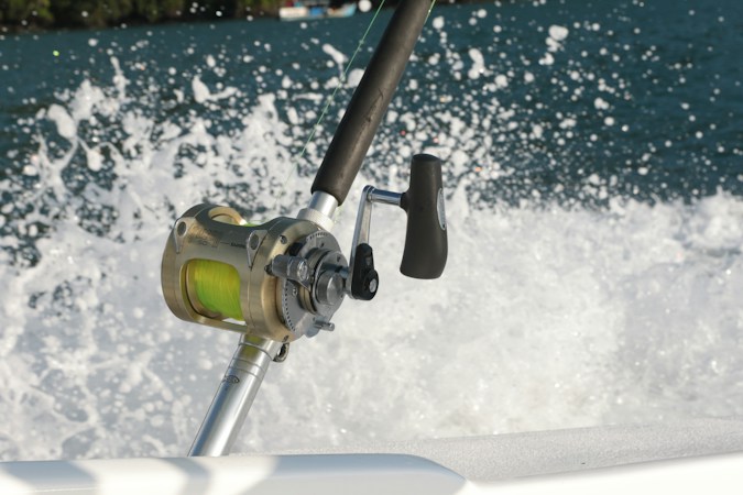 Gold conventional reel with yellow line on a rod in a boat's holder, white spray from the wake bursting behind it