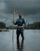 A man stands confidently in water, holding a golden trident. He is dressed in a shiny, textured superhero-like suit. The background is overcast with dark clouds, and there are trees and rocks providing a natural setting.