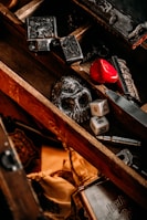 A wooden box contains various objects, including a black skull, two six-sided dice, a red heart-shaped object, and some metal items with intricate designs. The box has a rustic and vintage appearance with a mix of dark and warm tones.
