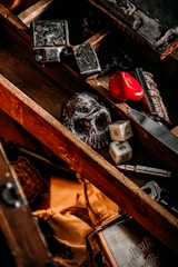 A wooden box contains various objects, including a black skull, two six-sided dice, a red heart-shaped object, and some metal items with intricate designs. The box has a rustic and vintage appearance with a mix of dark and warm tones.
