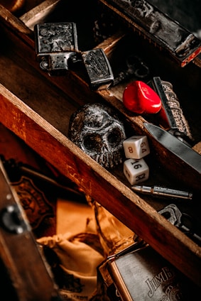 A wooden box contains various objects, including a black skull, two six-sided dice, a red heart-shaped object, and some metal items with intricate designs. The box has a rustic and vintage appearance with a mix of dark and warm tones.