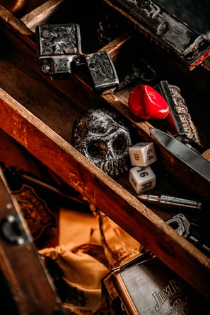 A wooden box contains various objects, including a black skull, two six-sided dice, a red heart-shaped object, and some metal items with intricate designs. The box has a rustic and vintage appearance with a mix of dark and warm tones.