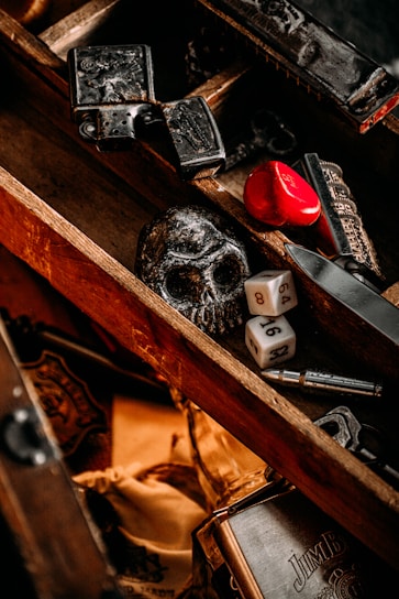 A wooden box contains various objects, including a black skull, two six-sided dice, a red heart-shaped object, and some metal items with intricate designs. The box has a rustic and vintage appearance with a mix of dark and warm tones.