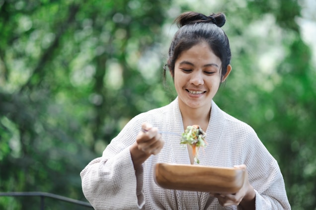 A person wearing a white robe is smiling while holding a wooden bowl and a fork with salad. The background is lush green, suggesting a natural outdoor setting.