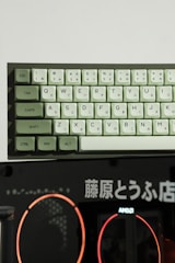 A mechanical keyboard with Japanese hiragana and standard QWERTY keys is positioned above a dark background featuring Japanese text and a logo for AMD. The keyboard has light green and white keys, with modifiers like ESC, TAB, and CTRL in green.