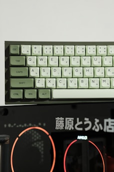 A mechanical keyboard with Japanese hiragana and standard QWERTY keys is positioned above a dark background featuring Japanese text and a logo for AMD. The keyboard has light green and white keys, with modifiers like ESC, TAB, and CTRL in green.
