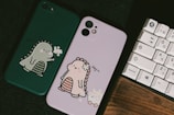 Two phone cases are displayed, one is green with an illustration of a cute dinosaur holding flowers, and the other is pink with a similar dinosaur design wearing a flower crown. A keyboard with Japanese characters sits beside them on a dark surface.