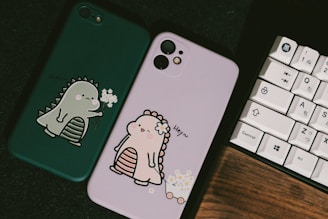 Two phone cases are displayed, one is green with an illustration of a cute dinosaur holding flowers, and the other is pink with a similar dinosaur design wearing a flower crown. A keyboard with Japanese characters sits beside them on a dark surface.