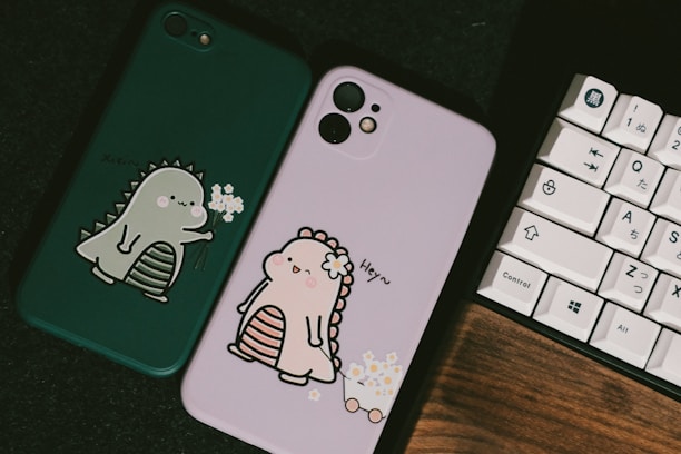 Two phone cases are displayed, one is green with an illustration of a cute dinosaur holding flowers, and the other is pink with a similar dinosaur design wearing a flower crown. A keyboard with Japanese characters sits beside them on a dark surface.
