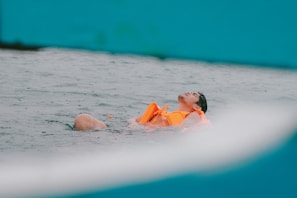 A person wearing an orange life vest is floating calmly on the water. The individual appears to be relaxed with their eyes closed, surrounded by rippling waves. The scene conveys a sense of tranquility and safety.