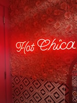 A neon sign displaying the words 'Hot Chica' is mounted on a wall covered with decorative roses. Below, geometric black and white tiles create a contrasting pattern, while decorative tiles with intricate designs are visible on the right edge. The scene is bathed in a warm red glow, enhancing the floral textures.