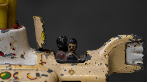 A close-up of a vintage, weathered toy vehicle with peeling paint. Two small, stylized figures are seated inside, featuring dark hair and simplistic details. The toy appears to be made of metal, showing signs of age and rust.
