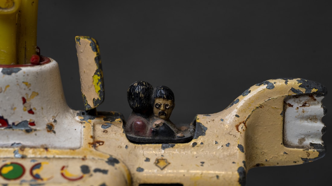 A close-up of a vintage, weathered toy vehicle with peeling paint. Two small, stylized figures are seated inside, featuring dark hair and simplistic details. The toy appears to be made of metal, showing signs of age and rust.