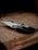 A folding knife with a sharp blade rests on a wooden surface, surrounded by soft shadows that give a rustic and slightly mysterious ambiance.