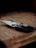A folding knife with a sharp blade rests on a wooden surface, surrounded by soft shadows that give a rustic and slightly mysterious ambiance.