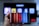 A hand holding a smartphone displaying the Netflix logo on its screen with a background of colorful vertical lines. In the background, a television screen shows blurry icons or profiles with various colors.