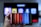 A hand holding a smartphone displaying the Netflix logo on its screen with a background of colorful vertical lines. In the background, a television screen shows blurry icons or profiles with various colors.