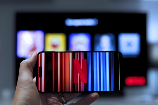 A hand holding a smartphone displaying the Netflix logo on its screen with a background of colorful vertical lines. In the background, a television screen shows blurry icons or profiles with various colors.