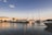 A serene marina filled with several luxurious yachts docked along the waterfront, with a clear sky above and calm waters reflecting the sunlight. Buildings with a Mediterranean architectural style stand in the background.