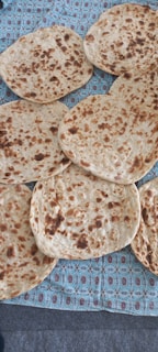 Several pieces of round, flat bread with a golden-brown surface and patches of darker spots are laid out on a patterned fabric background.