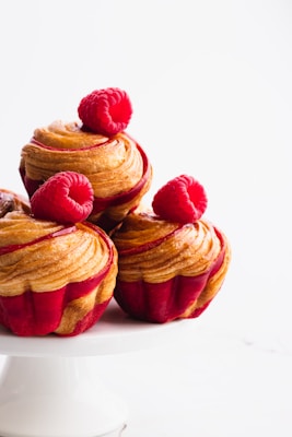 Delicately crafted pastries topped with fresh raspberries rest on a simple white cake stand. The pastries have a rich, golden-brown swirl with hints of red, giving them a freshly baked appearance.