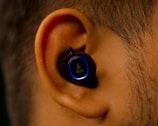 A close-up view of an ear with a black wireless earbud inserted. The earbud has a glowing blue ring around its surface, contrasting against the ear's natural skin tone.