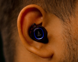 A close-up view of an ear with a black wireless earbud inserted. The earbud has a glowing blue ring around its surface, contrasting against the ear's natural skin tone.